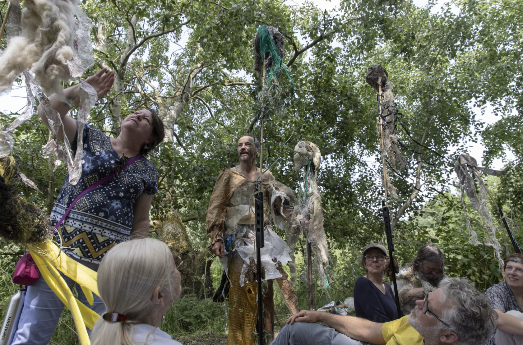 Seven people, most sat on the floor outdoors surrounded by green trees looking at materials attached to stakes.